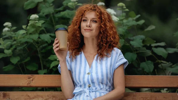 Happy redhead woman in dress holding paper cup while sitting on bench in park — Stock Photo