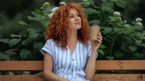 Cheerful redhead woman in dress holding paper cup while sitting on bench in park — Stock Photo