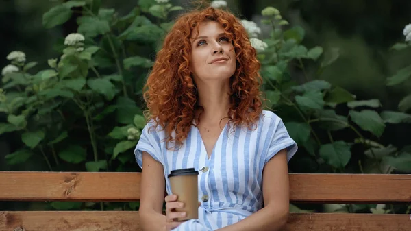 Positive redhead woman in dress holding paper cup while sitting on bench in park — Stock Photo