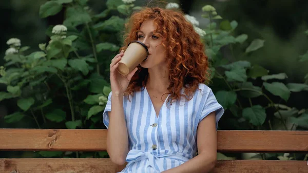 Encaracolado ruiva mulher no vestido beber café para ir enquanto sentado no banco no parque — Fotografia de Stock