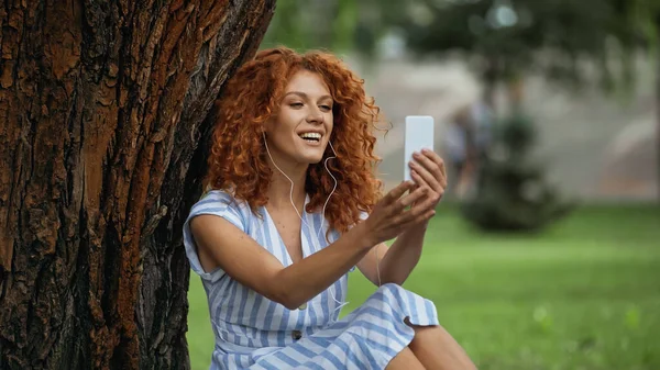 Happy redhead woman in blue dress and earphones listening music while looking at smartphone — Stock Photo