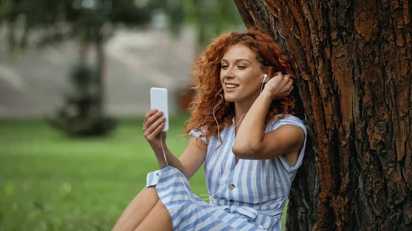 Mujer pelirroja satisfecha en vestido azul y auriculares escuchando música mientras mira el teléfono inteligente - foto de stock