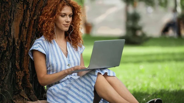 Curly redhead woman in dress listening music while using laptop in park — Stock Photo