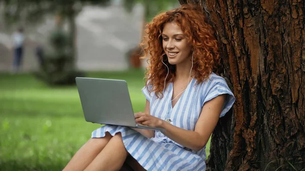 Mujer pelirroja feliz en vestido escuchando música mientras se utiliza el ordenador portátil en el parque - foto de stock