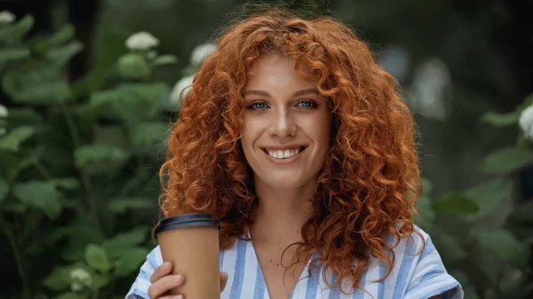 Cheerful redhead woman holding paper cup outdoors — Stock Photo