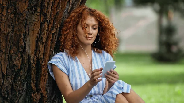 Mujer pelirroja feliz en vestido azul y auriculares escuchando música mientras usa el teléfono inteligente cerca del tronco del árbol - foto de stock