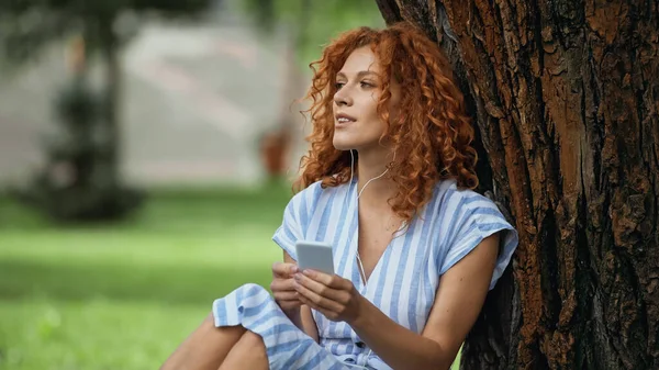 Mujer pelirroja complacida en vestido azul y auriculares escuchando música mientras usa el teléfono inteligente cerca del tronco del árbol - foto de stock