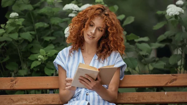 Curly redhead woman smiling while reading book and sitting on wooden bench in park — Stock Photo