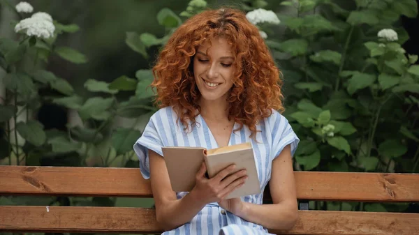 Feliz pelirroja sonriendo mientras lee libro y sentado en banco de madera en el parque - foto de stock