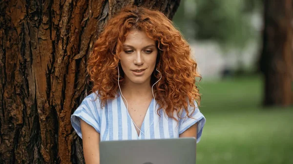 Mujer pelirroja joven escuchando música en los auriculares mientras usa el ordenador portátil en el parque - foto de stock