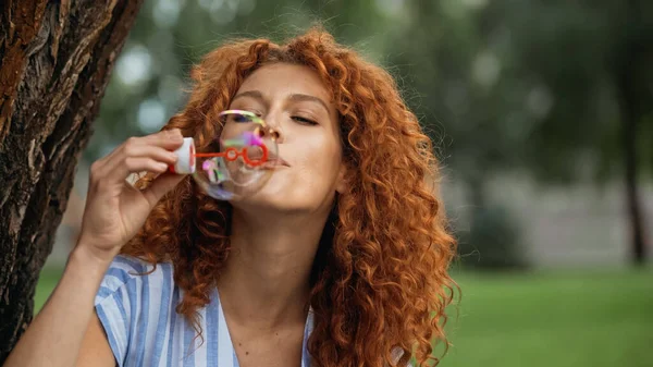 Pelirroja rizada soplando burbuja de jabón en el parque - foto de stock