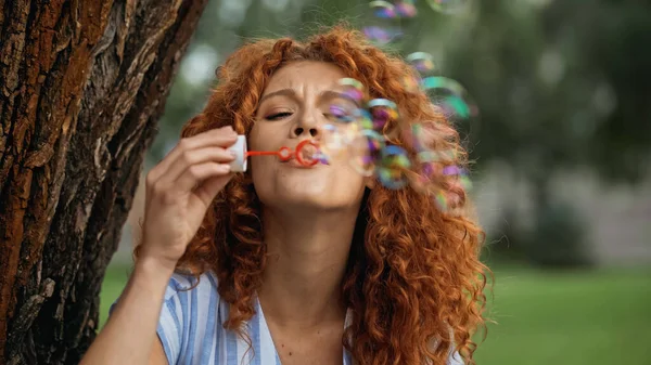 Curly redhead woman frowning while blowing soap bubbles in park — Stock Photo