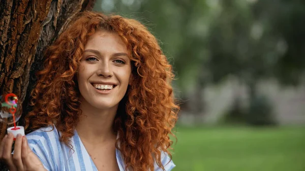 Glückliche rothaarige Frau hält Zauberstab mit Seifenblase im Park — Stockfoto