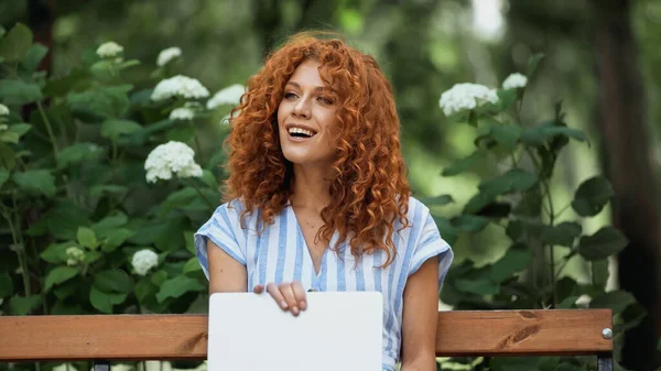 Mujer pelirroja feliz usando el ordenador portátil mientras está sentado en el banco en el parque - foto de stock