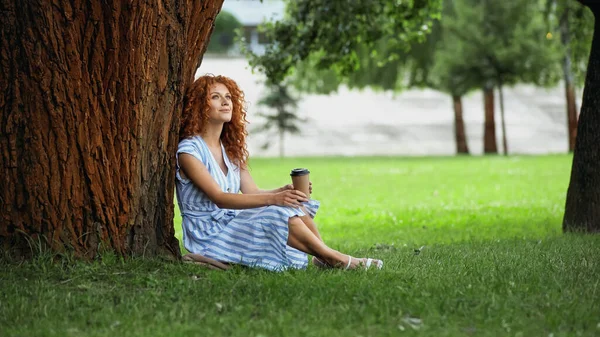 Piena lunghezza della donna rossa sognante in abito seduto sotto il tronco d'albero e tenendo tazza di carta — Foto stock