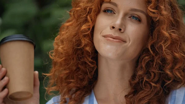 Heureuse rousse femme avec les yeux bleus tenant tasse en papier à l'extérieur — Photo de stock