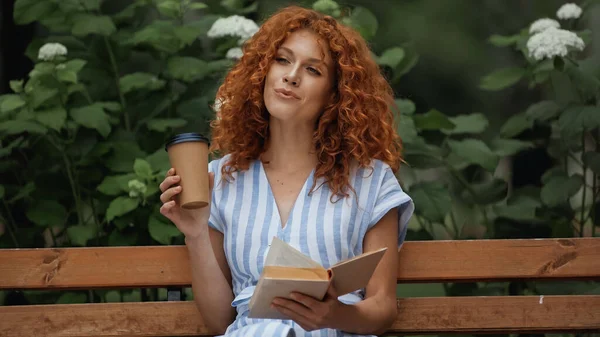 Erfreut rothaarige Frau im gestreiften Kleid hält Buch und Pappbecher in der Hand, während sie auf einer Bank im Park sitzt — Stockfoto