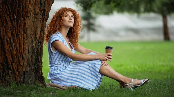 Dreamy redhead woman in blue dress sitting under tree trunk and holding coffee to go — Stock Photo