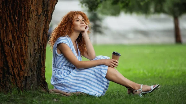 Dreamy redhead woman in blue dress smiling while sitting under tree trunk and holding coffee to go — Stock Photo