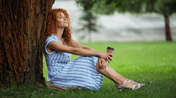 Smiling redhead woman in blue dress sitting under tree trunk and holding coffee to go — Stock Photo