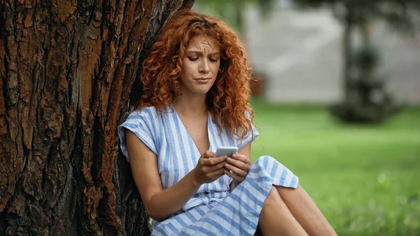 Upset redhead woman in blue dress sitting under tree trunk and using smartphone — Stock Photo