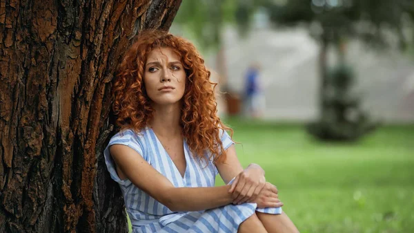 Upset redhead woman in blue dress sitting under tree trunk in park — Stock Photo