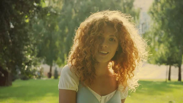 Sol en el pelo rojo rizado de la mujer sonriendo mientras mira a la cámara en el parque - foto de stock