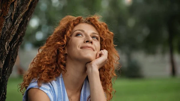 Mujer soñadora con el pelo rizado mirando hacia arriba y sonriendo en el parque - foto de stock