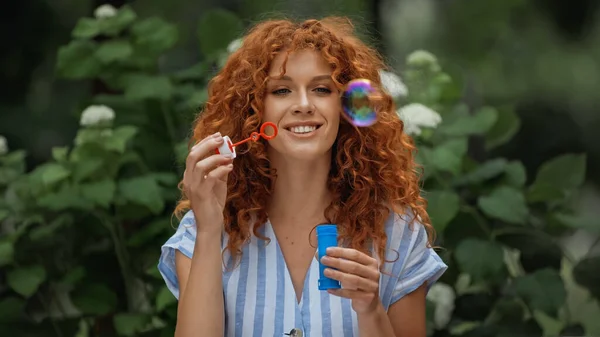 Cheerful redhead woman looking at blurred soap bubbles in park — Stock Photo