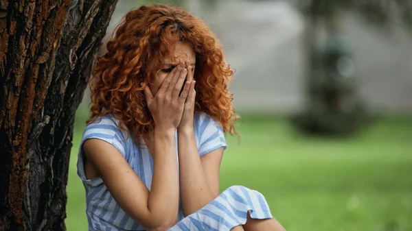 Jovem triste com rosto de cobertura de cabelo vermelho enquanto chorava no parque — Fotografia de Stock