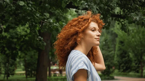 Кудрявая молодая женщина с рыжими волосами стоит в зеленом парке — стоковое фото