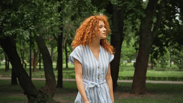 Красивая молодая женщина с рыжими волосами, стоящая в синем полосатом платье в зеленом парке — стоковое фото