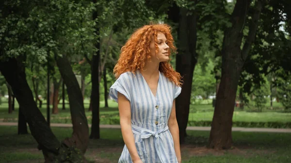 Bonita mujer joven con el pelo rojo de pie en vestido de rayas azules en el parque verde - foto de stock