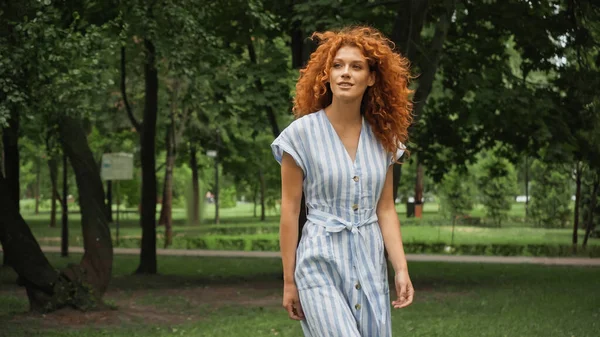 Joyful young woman with red hair standing in blue striped dress in green park — Stock Photo