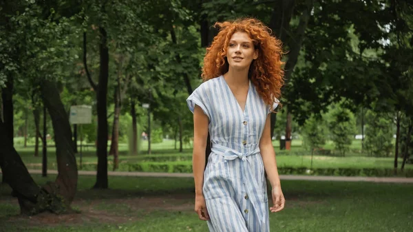 Alegre joven con el pelo rojo de pie en vestido de rayas azules en el parque verde - foto de stock