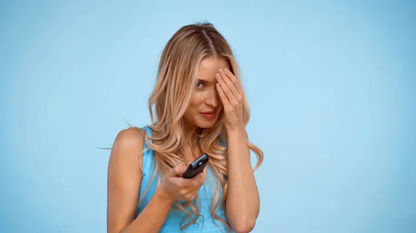 Mujer rubia sosteniendo mando a distancia y cubriendo la cara aislada en azul - foto de stock