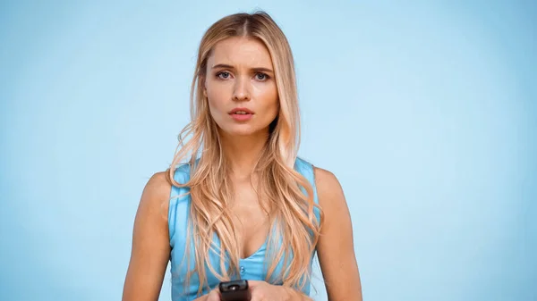 Upset blonde woman holding remote controller and looking at camera isolated on blue — Stock Photo