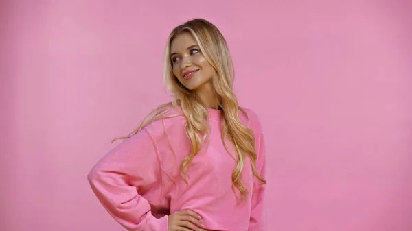 Smiling woman in sweatshirt holding hand on hip isolated on pink — Stock Photo