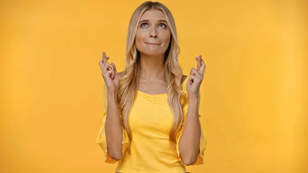 Mujer rubia cruzando los dedos y mirando hacia arriba aislado en amarillo - foto de stock