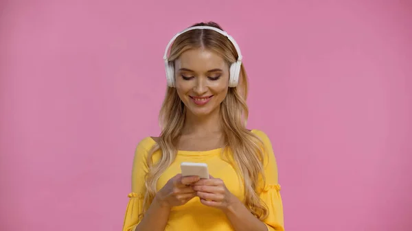 Mulher loira positiva em fones de ouvido usando smartphone isolado em rosa — Fotografia de Stock