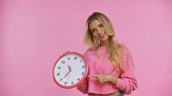 Lächelnde blonde Frau zeigt mit dem Finger auf Uhr in rosa — Stockfoto