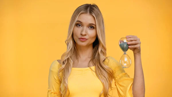 Blonde woman looking at camera while holding hourglass isolated on yellow — Stock Photo