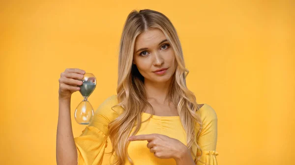 Blonde woman pointing at hourglass with finger isolated on yellow — Stock Photo