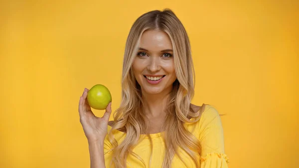 Mulher loira positiva olhando para a câmera e segurando maçã isolada no amarelo — Fotografia de Stock