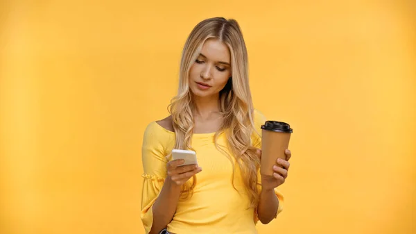 Mujer rubia usando smartphone y sosteniendo taza de papel aislada en amarillo - foto de stock