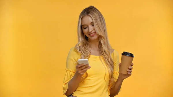 Positivo donna bionda utilizzando smartphone e tenendo il caffè per andare isolato sul giallo — Foto stock