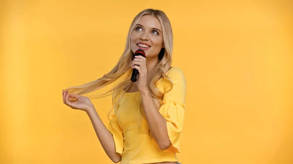 Позитивная блондинка поет караоке и трогает волосы, изолированные на желтом — стоковое фото