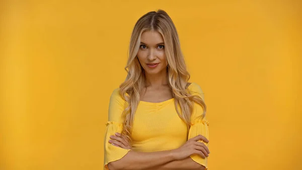 Pretty blonde woman smiling at camera and crossing arms isolated on yellow — Stock Photo