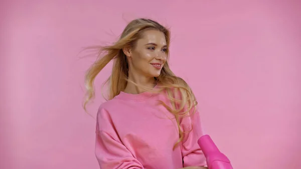 Smiling blonde woman using hair dryer isolated on pink — Stock Photo