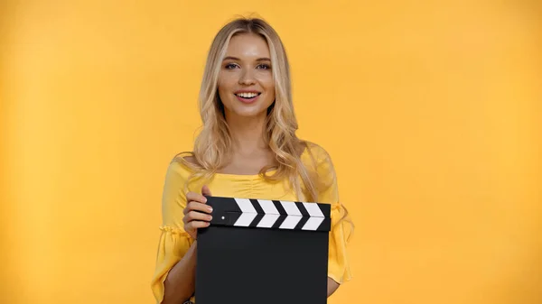 Mujer rubia feliz sosteniendo el clapboard y mirando a la cámara aislada en amarillo - foto de stock